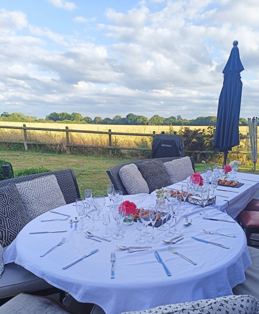 A garden table set ready for dinner with a whilte tablecloth and flowers.  With views over fields and a blue sky with fluffy clouds. 